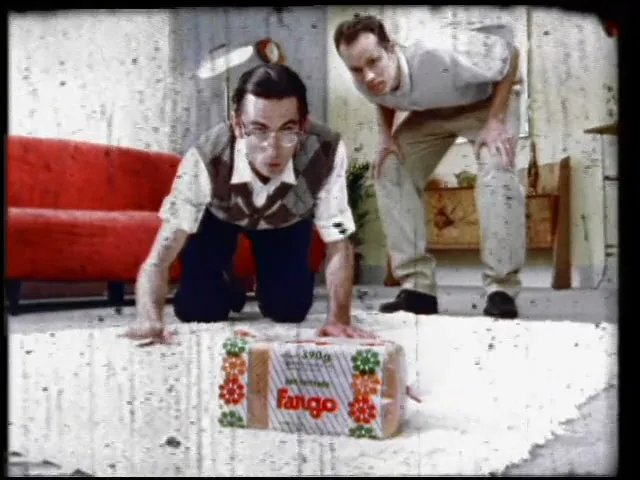 Two men, one on his knees and one standing, look intently at a package of Fargo bread on a white rug in a living room.