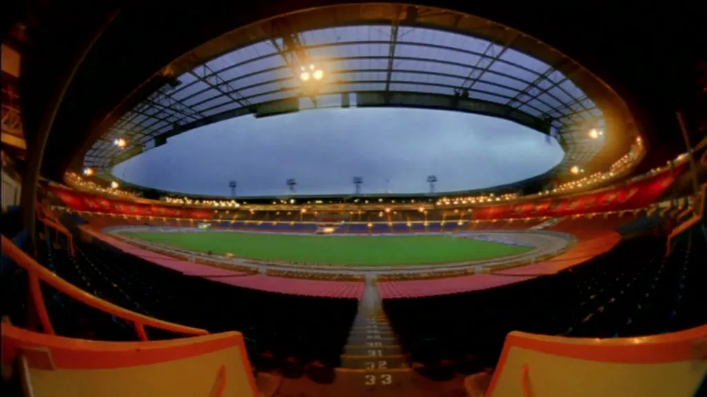 A wide-angle shot of an empty football stadium with rows of seats and a green field under a cloudy sky, as the lights turn on.