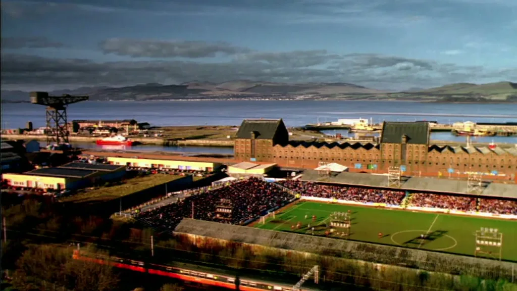 An aerial view of a football stadium full of spectators, with a harbor, industrial buildings, and mountains in the background under a cloudy sky.