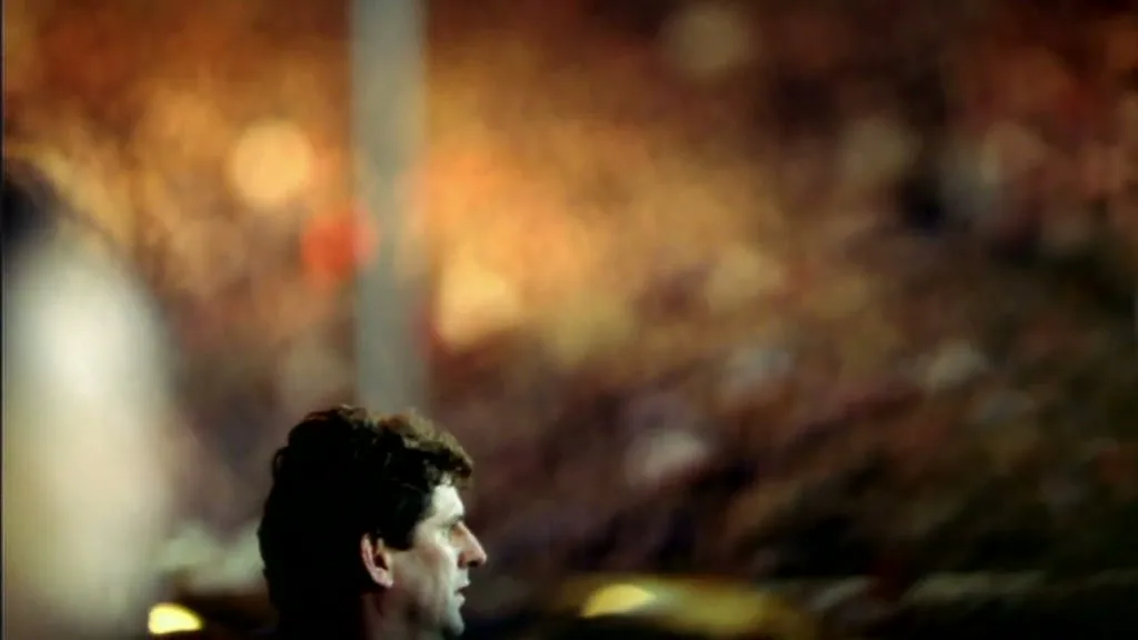 A man with dark curly hair looks right, with a blurred background of stadium lights and crowd, at night.