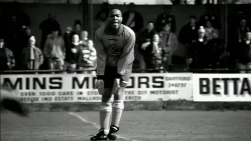A male soccer player in a light-colored uniform stands on a field, looking distressed, with other players and spectators blurred in the background.