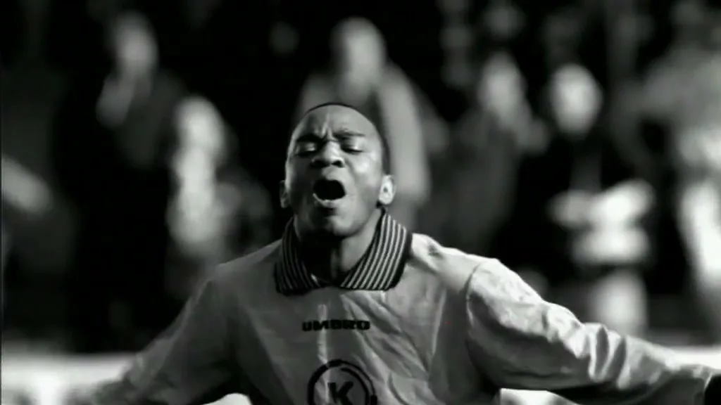 A male soccer player in a light-colored jersey with dark stripes on the collar screams in agony on a field, with blurred spectators in the background.