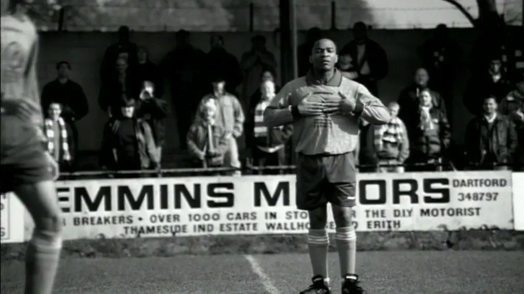 A male soccer player in a light jersey and dark shorts stands on a field, holding his chest, as another player walks past him in front of a crowd and a billboard.