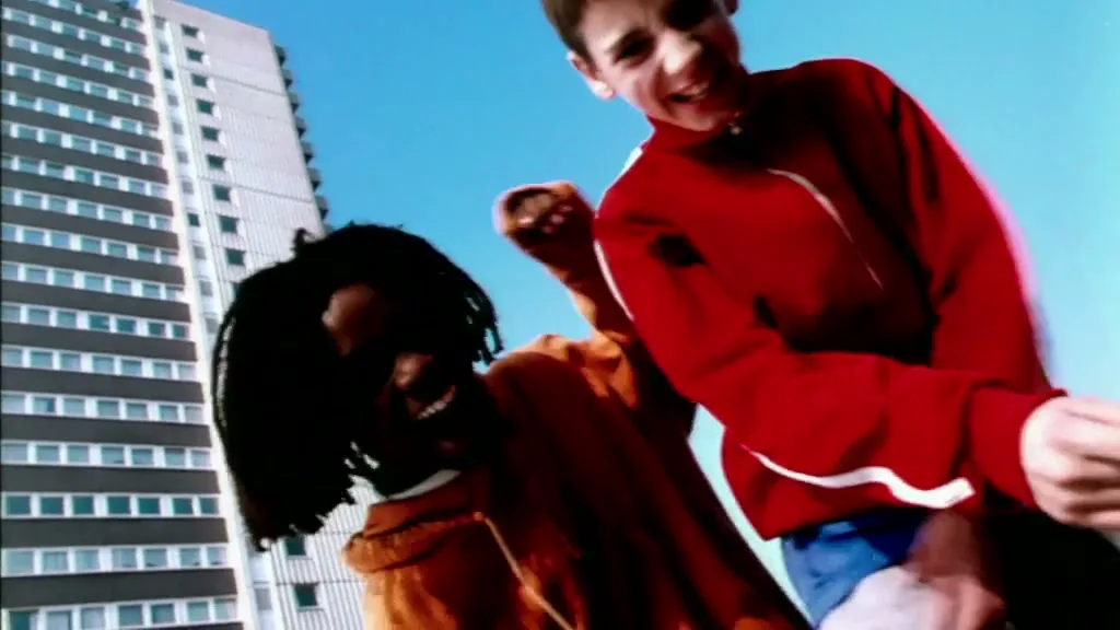 Two young boys, one in a red jacket and one in an orange hoodie, laugh and point at the camera in front of a tall apartment building on a sunny day.