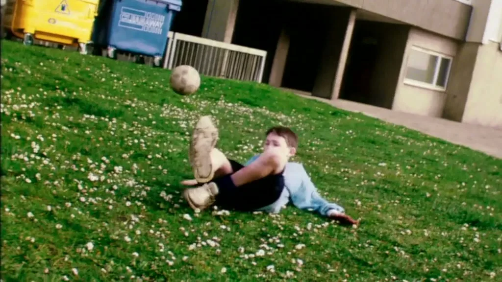 A young boy in a light blue shirt and dark shorts falls backward onto a grassy hill with a soccer ball nearby, in front of apartment buildings and trash bins.