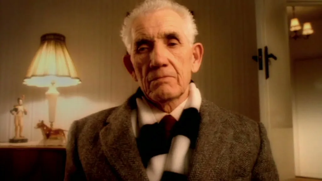 An elderly man with white hair and a black and white scarf looks directly at the camera in a dimly lit room.