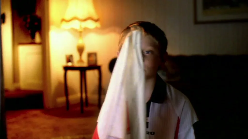 A young boy in an England football shirt looks sad, then raises a small England flag to his face, then lowers it, in a dimly lit living room.