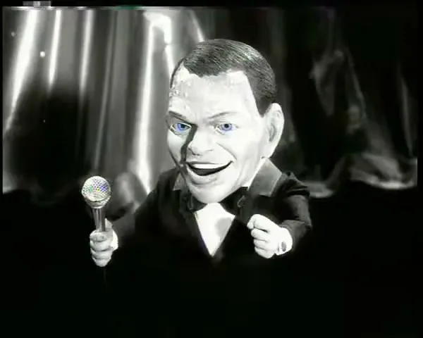 A puppet of Frank Sinatra, with blue eyes, sings into a microphone on a stage with a curtain backdrop.