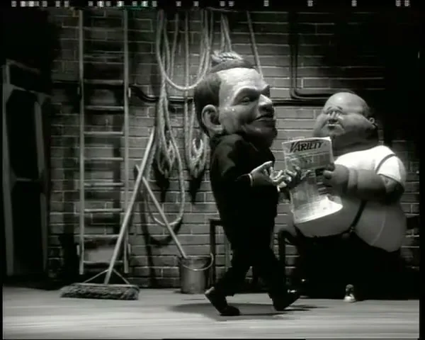 A puppet with a large head resembling Frank Sinatra walks past a film set, followed by a shorter, stout puppet holding a newspaper, then a taller puppet, all in black and white.