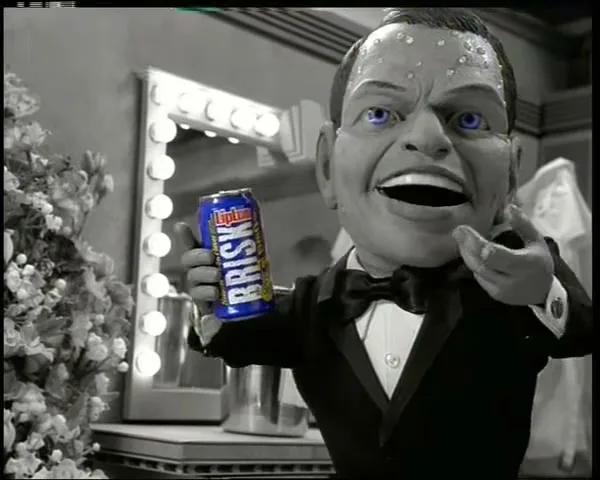 A puppet of Frank Sinatra with a large head, wearing a tuxedo, holds a can of Lipton Brisk iced tea in a dressing room.
