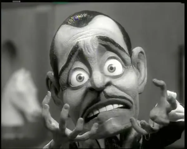 A stop-motion puppet of a man with a mustache and wide eyes looks distressed, raising his hands in a black and white office setting.