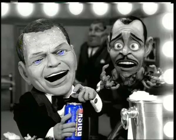Two puppet caricatures of famous male singers, one holding a blue Brisk can, are in a dressing room with a vanity mirror.