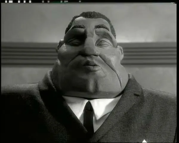 A claymation man in a suit looks surprised, with his eyes wide open and mouth agape, in an interior setting.