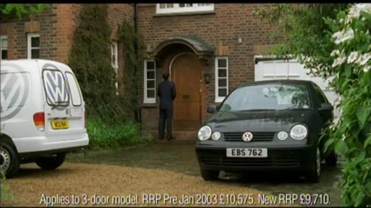 A man in a suit stands at the front door of a brick house, while a black Volkswagen Polo and a white Volkswagen van are parked in the driveway.
