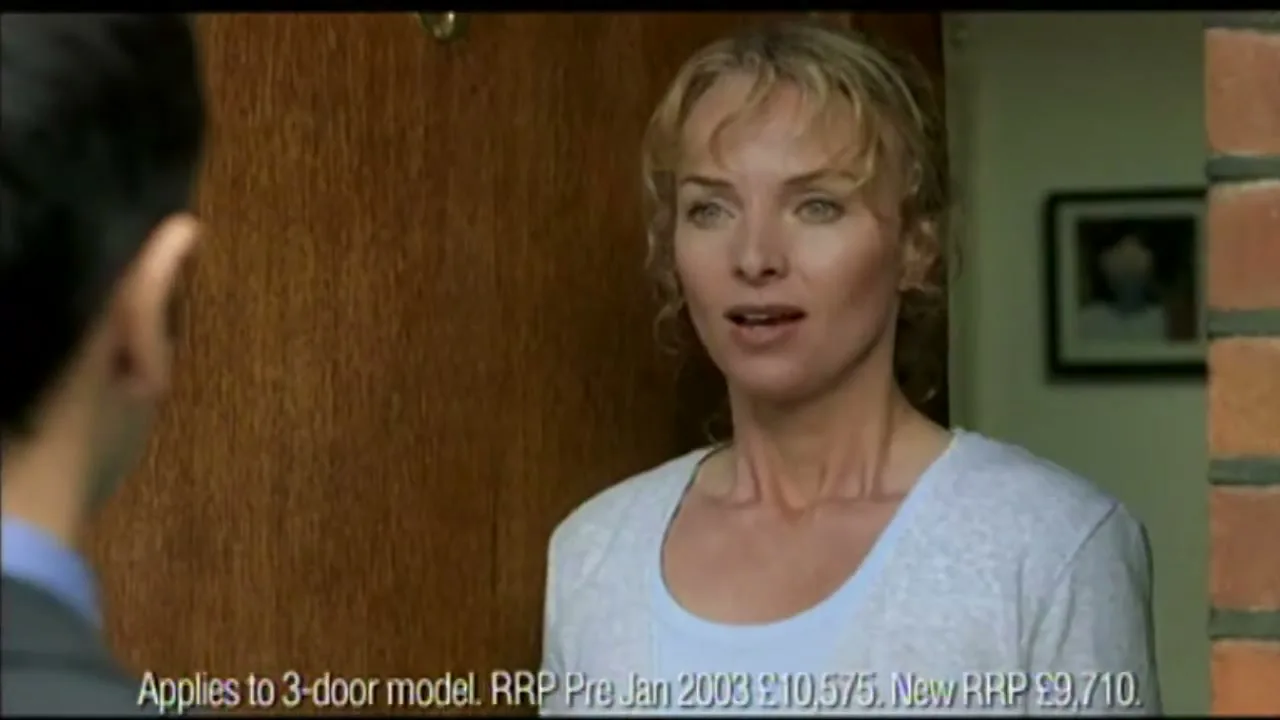 A blonde woman in a white top opens the door to a man in a suit, speaking to him at the entrance of her home.