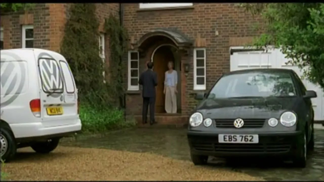 A man in a blue jacket runs past a black Volkswagen car and a white Volkswagen van parked in front of a brick house, where a man and woman stand in the doorway.