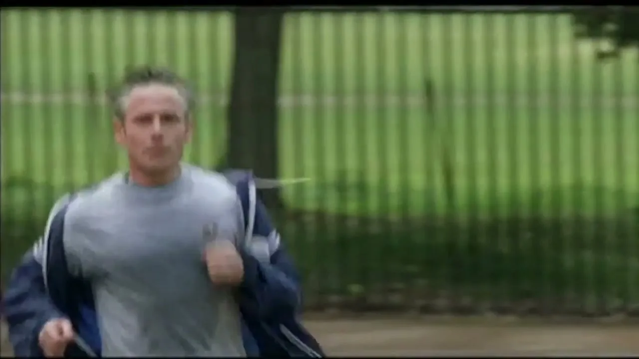 A man in his 30s or 40s, wearing a grey t-shirt and blue jacket, jogs through a park on a bright day.