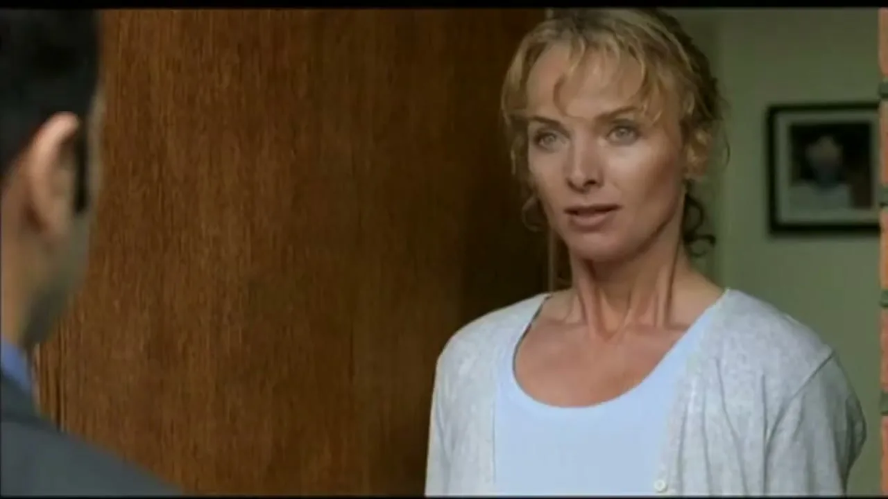 A blonde woman in her 40s, wearing a light blue top and grey cardigan, stands in a doorway talking to a man whose back is to the camera.