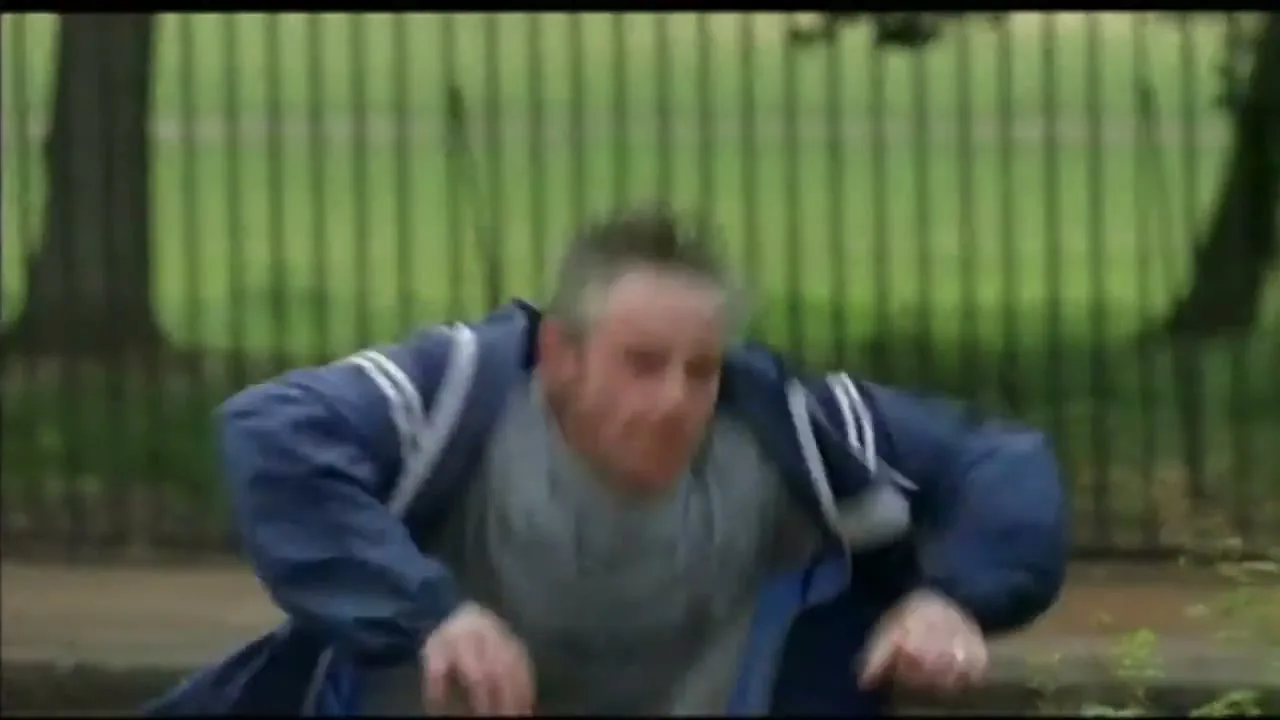 A middle-aged man in athletic wear jogs on a sidewalk next to a green fence, then suddenly falls forward.