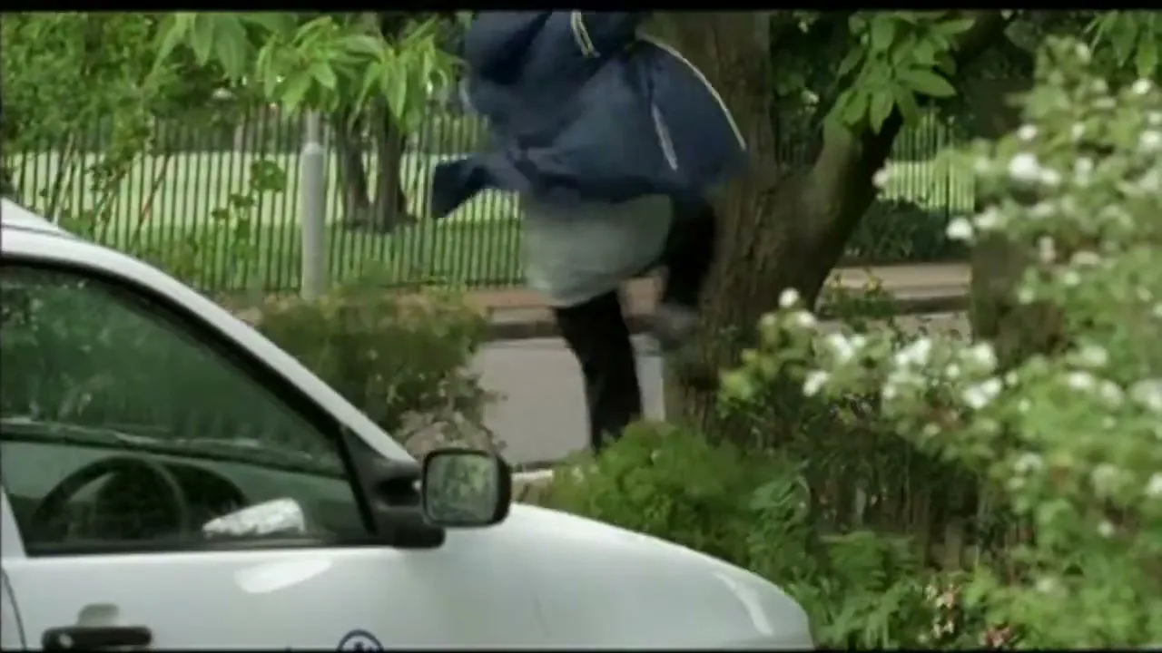 A man in a blue jacket and grey shirt jumps over a hedge next to a white car on a suburban street during the day.