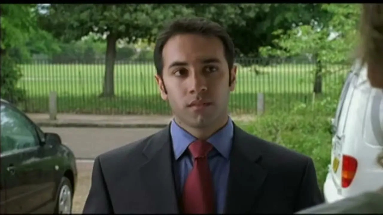 A man in a suit looks at someone off-camera, standing outdoors with cars and trees in the background.