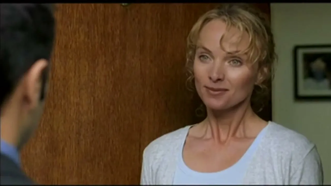 A blonde woman in a light blue top and grey cardigan smiles at a man, standing in a doorway in an interior setting.