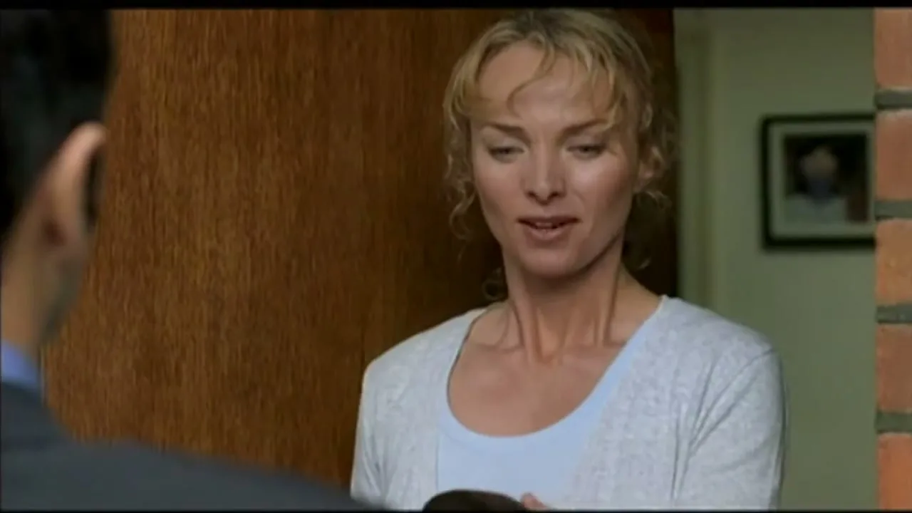 A blonde woman in a light blue top and white cardigan stands in a doorway, looking at a man whose back is to the camera, and smiles as she receives something.