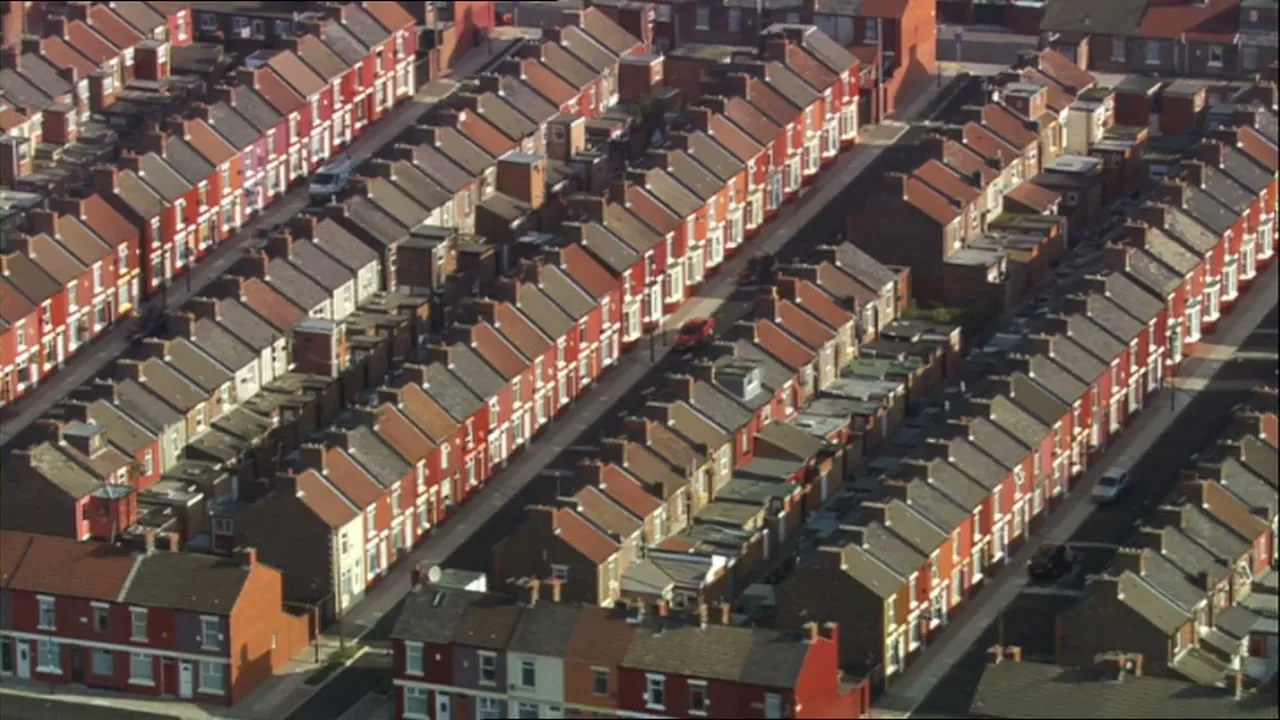 An aerial shot shows rows of terraced houses with red roofs and small backyards in a dense urban neighborhood.
