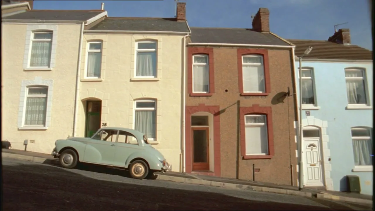 A light green vintage car is parked on a steep residential street with colorful terraced houses under a clear sky.