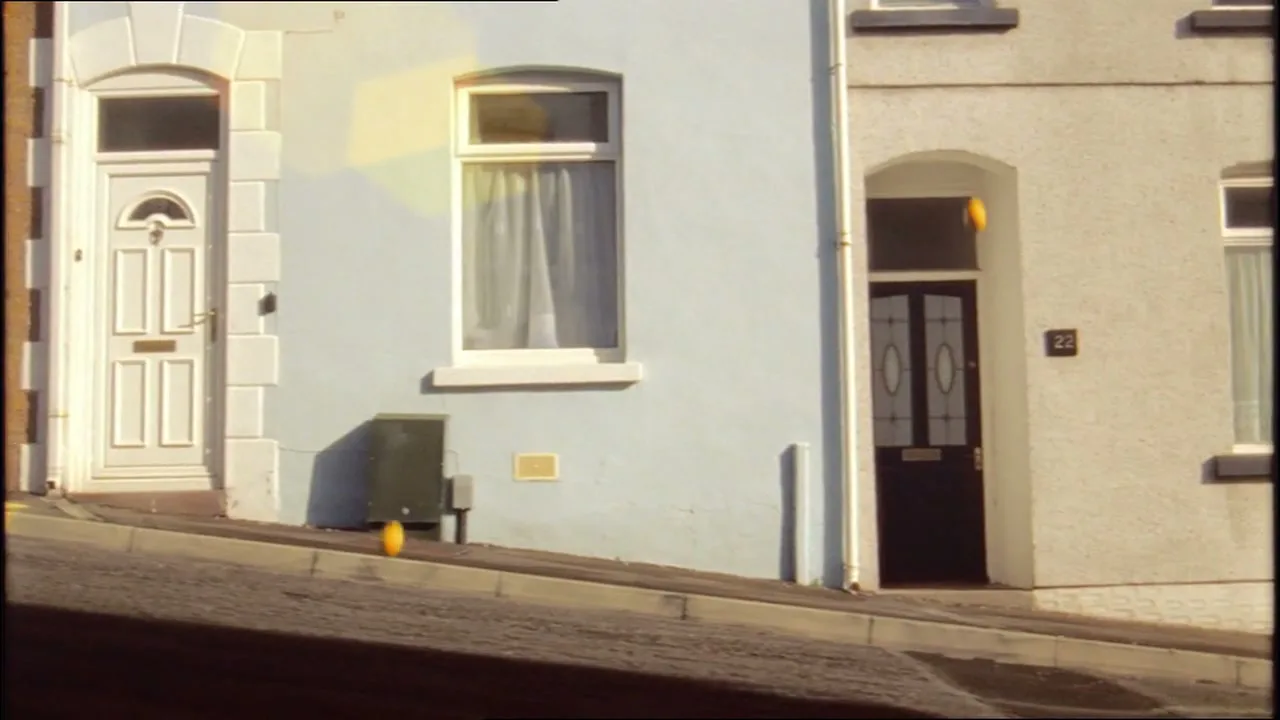 A small orange fruit rolls down a sloped sidewalk in front of two houses, one light blue and one white, on a sunny day.