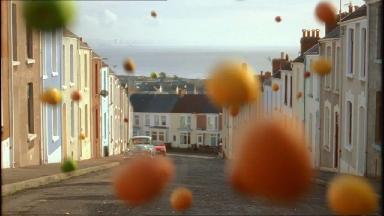 A wide shot of a residential street with colorful houses, as various fruits fall from the sky and bounce on the road, with the sea in the background.