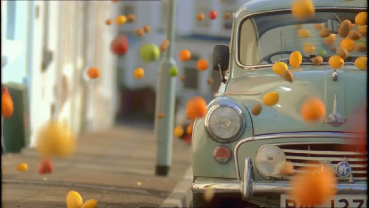 A vintage car is parked on a residential street as various fruits, including oranges and lemons, fall and bounce around it.