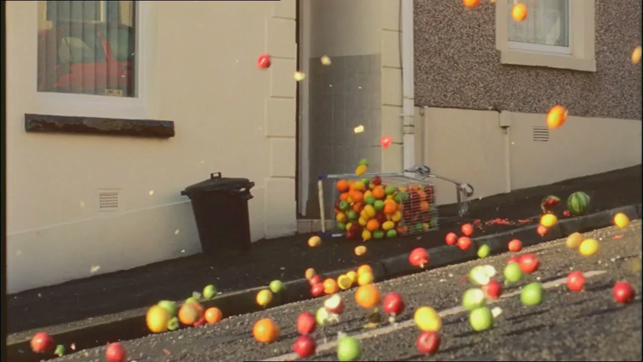 A shopping cart full of fruit tumbles down a residential street, spilling its contents everywhere as the fruit bounces and rolls.