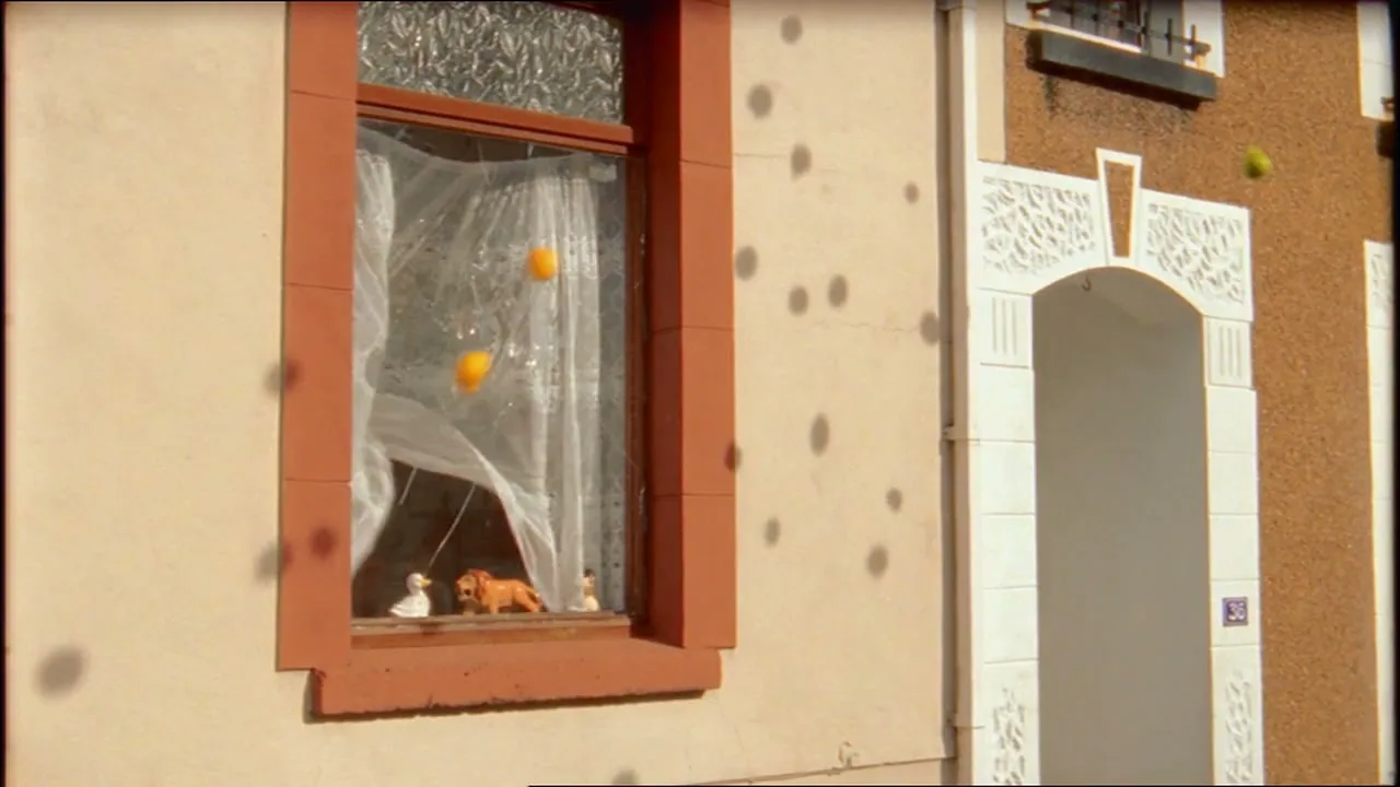 A window with orange trim and white curtains is shown on the side of a building as small, colorful fruit-like objects are thrown at it, causing the glass to shatter.