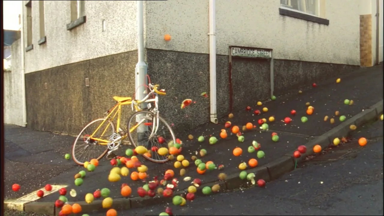 A yellow bicycle is parked next to a lamppost on a street corner as a cascade of colorful fruit tumbles down a hill.