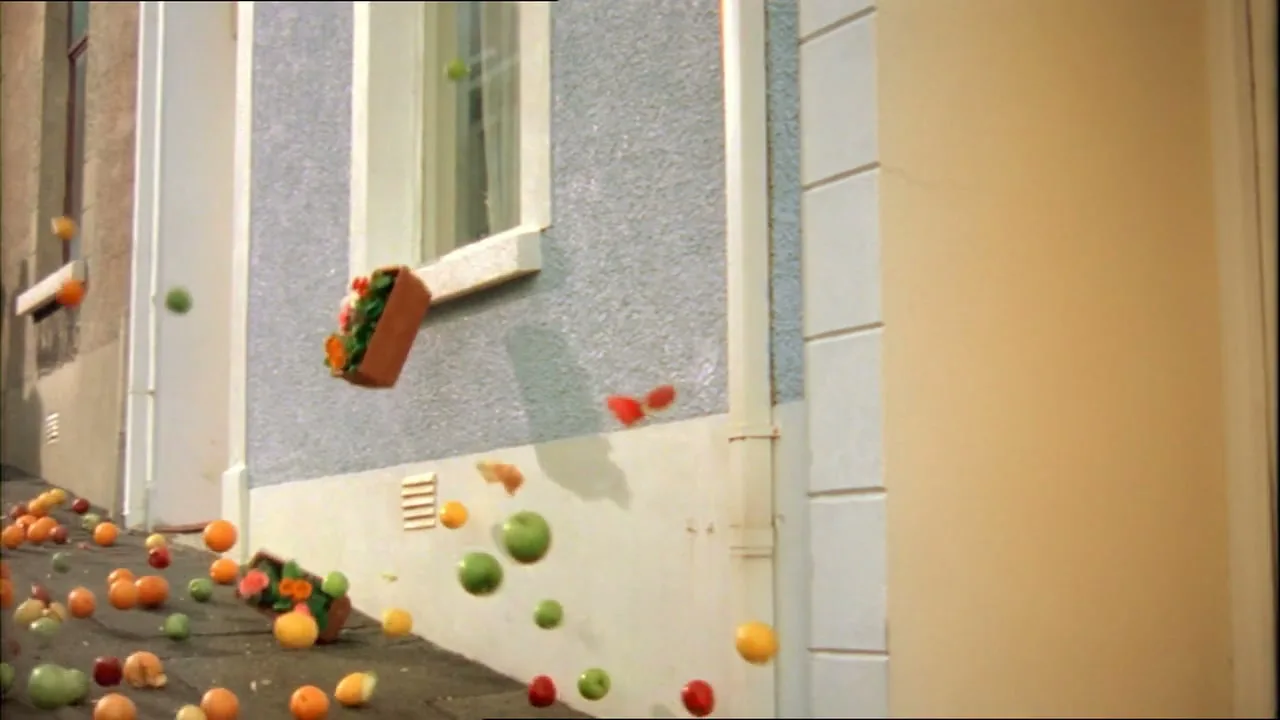 A window box falls from a window, spilling fruit onto the ground, as more fruit bounces and rolls down a sloped alleyway.