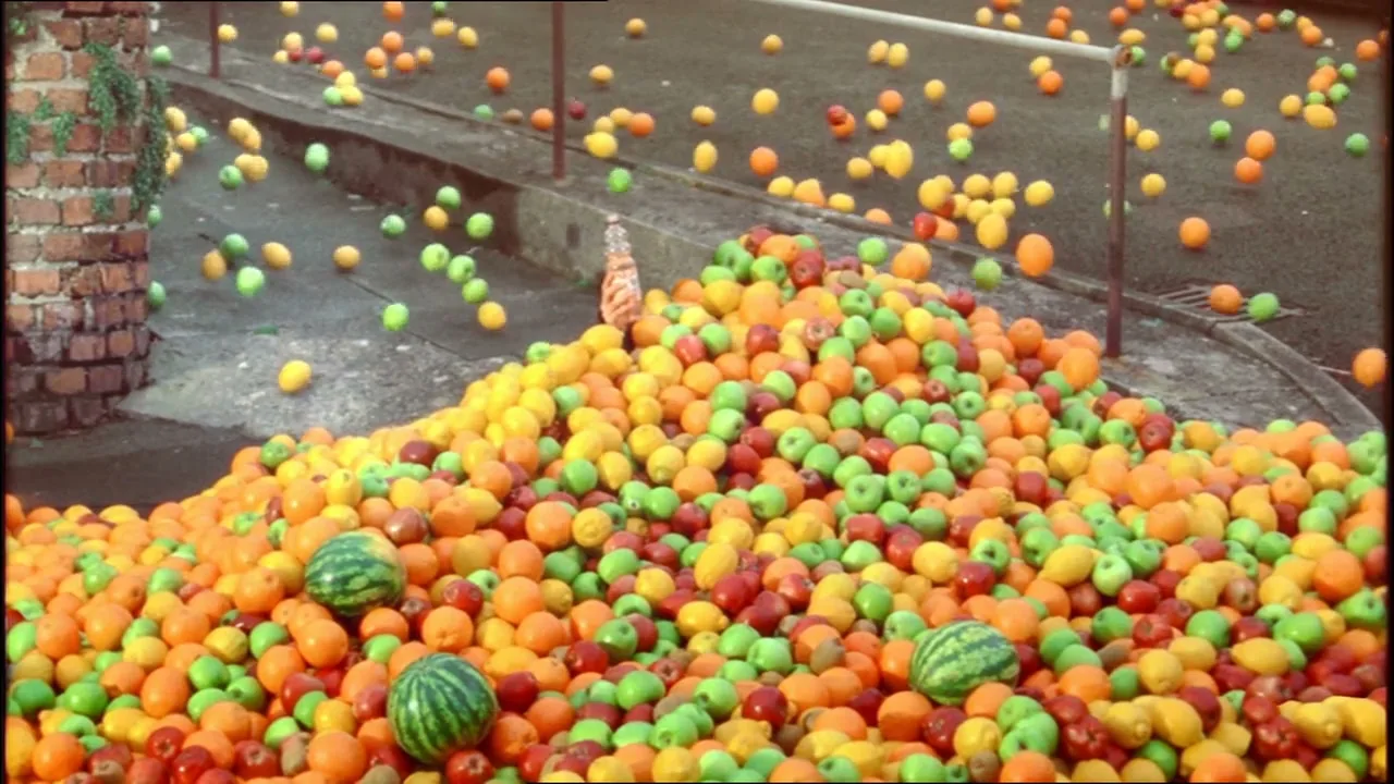A wide shot shows a massive pile of oranges, green apples, and watermelons on a paved street, with more fruit rolling down a slope.