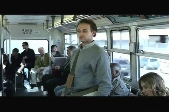 A young man in a grey sweater stands in a moving bus, looking around at other passengers.