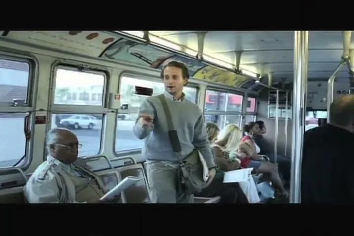 A young man in a grey sweater and messenger bag stands on a moving bus, pointing and talking to other passengers.