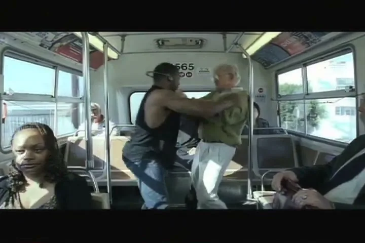 A young man in a black tank top and an older man in a green shirt are fighting on a bus, while other passengers look on.