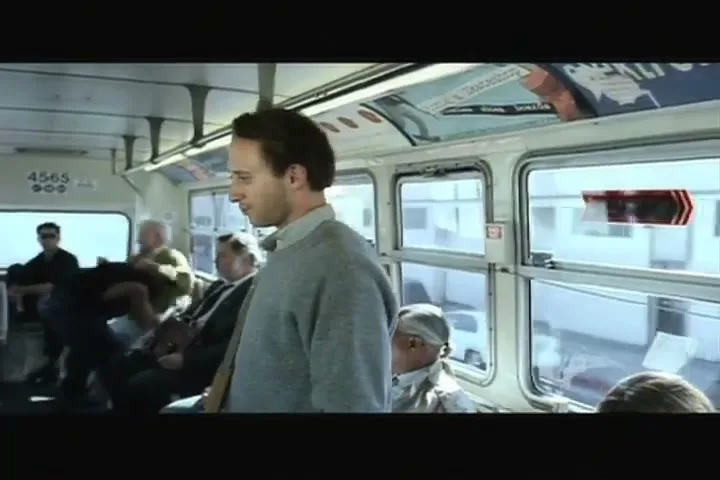 A young man in a grey sweater stands in a moving bus, looking to his right and gesturing with his hand, surrounded by other passengers.