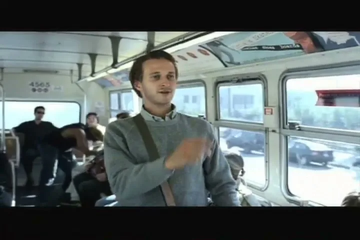 A young man in a grey sweater stands in a moving bus, looking around and gesturing with his hand as other passengers are visible in the background.