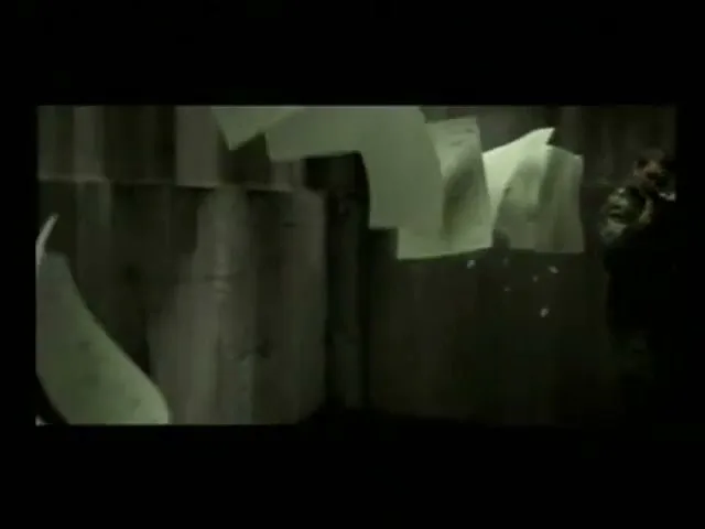 A person in dark clothing stands in a dark, industrial-looking space, holding a large, light-colored sheet of paper that appears to be hit by a bullet, causing more papers to fly around.