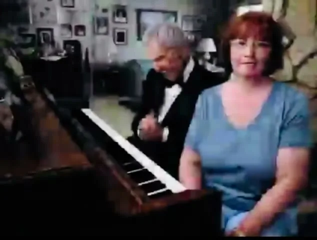A woman with red hair and glasses sits in the foreground, while a man in a tuxedo plays the piano in the background of a living room.