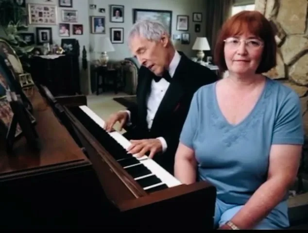 A woman with red hair and glasses sits in the foreground, looking at the camera, while a man in a tuxedo plays the piano behind her in a living room.