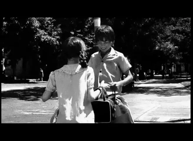 Two children, a boy and a girl, stand facing each other on a sunny day in a park, the boy holding a bag.