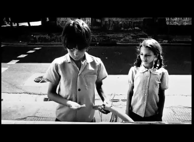 Two children, a boy and a girl, stand outdoors in a black and white shot. The boy holds a small object, while the girl watches him.