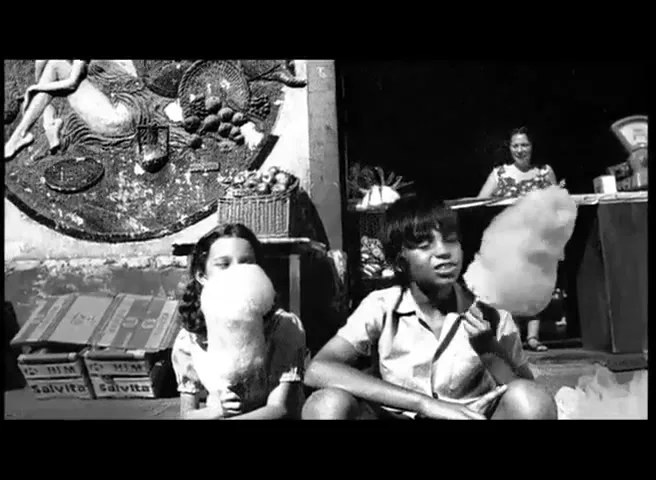 Two children, a boy and a girl, sit outdoors eating cotton candy, with a woman visible in the background, all in black and white.