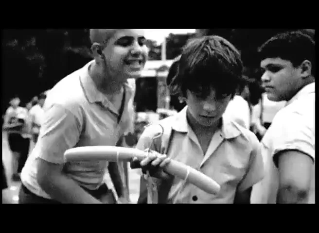 A young boy with dark hair holds a wooden toy, looking downcast, while other boys stand around him in a black and white outdoor setting.