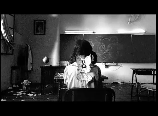 A young girl with pigtails and a mouse mask sits at a desk in a messy classroom, facing away from the camera, in black and white.